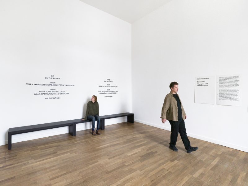 Ein Besucher geht rückwärts durch einen hellen Ausstellungsraum, während eine Frau auf einer Bank sitzt.